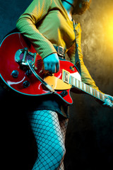 Close-up photo of young hipster woman legs and red guitar in neon lights. Rock musician is playing...