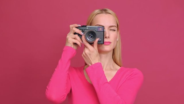 Attractive young woman blonde in a red T-shirt and jeans posing on a pink background. Photographs on a mirrorless camera in retro style and smiling. The concept of the photographer.