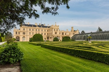 Fototapeta na wymiar Lednice, Czech Republic - May 28 2019: Famous Lednice castle in South Moravia with yellow facade. Garden with green lawn, bushes, trees and greenhouse. Sunny spring day, blue sky, white clouds.