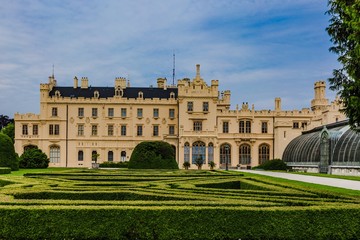 Obraz premium Lednice, Czech Republic - May 28 2019: Famous Lednice castle in South Moravia with yellow facade. Garden with green lawn, bushes, trees and greenhouse. Sunny spring day, blue sky, white clouds.