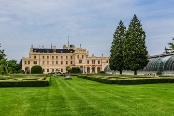 Fototapeta na wymiar Lednice, Czech Republic - May 28 2019: Famous Lednice castle in South Moravia with yellow facade. Garden with green lawn, bushes, trees and greenhouse. Sunny spring day, blue sky, white clouds.