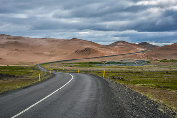Obraz premium Curvy road, empty meadow and red mountains in the background in Myvatn region, overcast day in summer