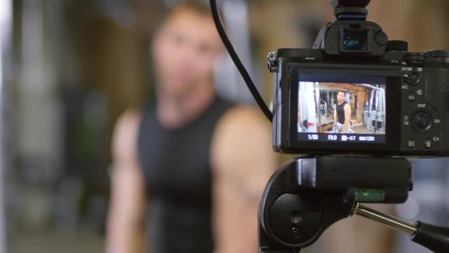 Rack focused shot of muscular male coach doing triceps cable pushdown and speaking on camera while recording his workout in gym