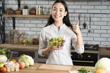 Front view of happy young woman holding glass of bowl with salad showing thumb up sign in kitchen