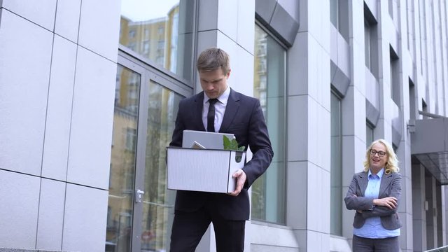 Sad male manager with stuff box looking gloating female boss, company dismissal