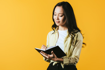 smiling girl writing in notebook with pen, isolated on yellow