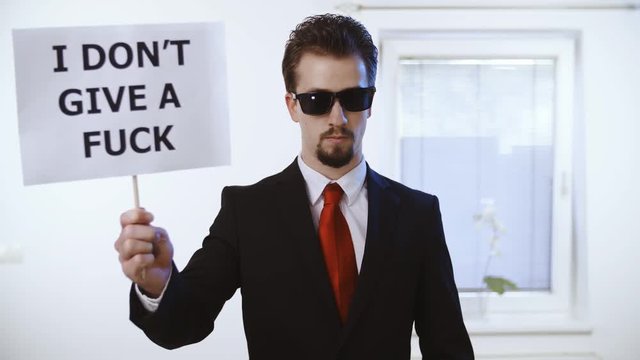 Man in business suit hold a sign I don't give a fuck 4K. Medium shot of a male person in focus dressed up nicely with a red tie. Wall with clock and window in the background.