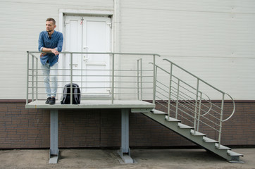 bearded young man in jeans and a denim shirt is standing on the porch of an industrial building near a closed door. firing concept