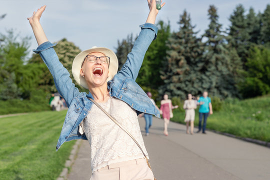 Happy Young Caucasian Bald Woman In Hat And Casual Clothes Enjoying Life After Surviving Breast Cancer. Portrait Of Beautiful Hairless Girl Smiling During Walk At City Park After Curing Disease