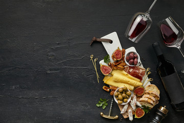 Obraz na płótnie Canvas Two glasses of red wine and a tasty cheese plate with fruit, grape, nuts and toasted bread on a wooden kitchen plate on the black stone background, top view, copy space. Gourmet food and drink.