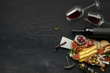 Obraz na płótnie Canvas Two glasses and bottle of red wine and a tasty cheese plate with fruits, olives and toasted bread on a wooden kitchen plate on the black stone background, top view, copy space. Gourmet food and drink.