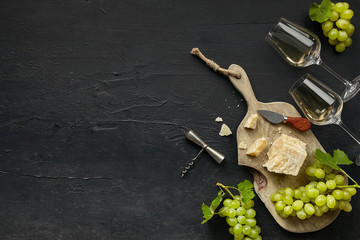 Two glasses of white wine and a tasty cheese plate with fruit on a wooden kitchen plate on the black stone background, top view, copy space. Gourmet food and drink.