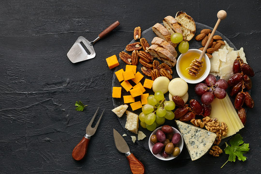 Top view of tasty cheese plate with fruit, grape, nuts and honey on a circle kitchen plate on the black stone background, top view, copy space. Gourmet food and drink.
