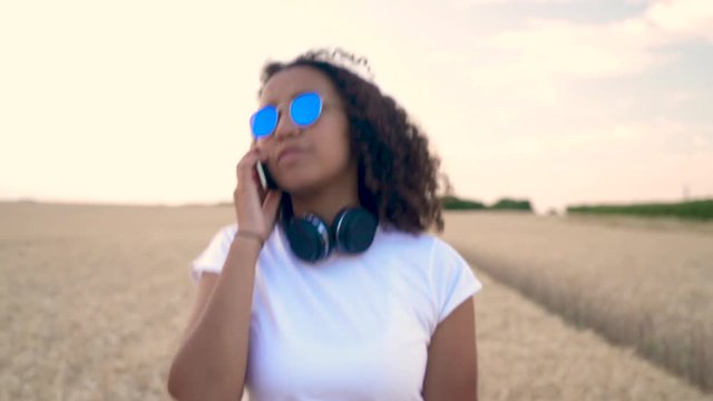 Slow motion follow shot video of beautiful biracial African American girl teenager young woman in white T-shirt and blue sunglasses walking listening to music on wireless headphones 