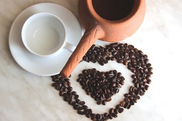 brown clay Turk for cooking Turkish coffee, white clean Cup and saucer and roasted coffee beans in the shape of a heart on the table, selective focus