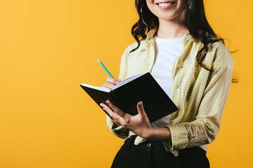 cropped view of smiling girl writing in notebook with pen, isolated on yellow