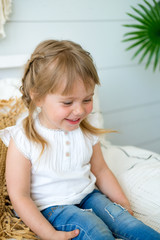 Fototapeta premium Beautiful little girl sitting on the bed in the bedroom. the baby has blonde hair and blue eyes. Jeans and white t-shirt