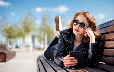 Woman sitting on bench among urban space and reading ebook using digital reader