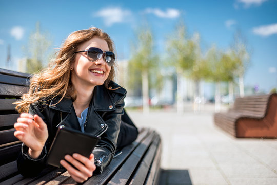 Woman sitting on bench among urban space and reading ebook using digital reader