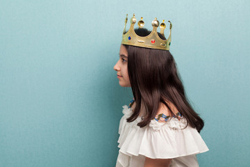 Side view portrait of serious beautiful teenager brunette girl wear golden crown symbol of princess in white dress standing and looking away. Indoor, isolated, studio shot, blue background © khosrork