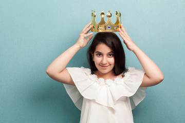 Portrait of young beautiful princess brunette girl in white dress standing and wear golden diadem crown with smile, looking at camera. Indoor, isolated, studio shot, blue background © khosrork