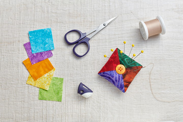 Pin cushion, scissors, spool of thread, thimble on cream-white wooden surface