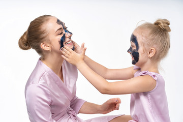 little daughter makes her mother a cosmetic mask on her face. Daughter cares, helps mom. Little daughter and young mother.