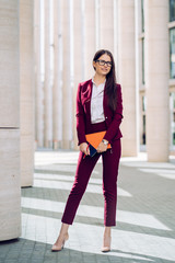 Obraz premium Stylish brunette business woman in maroon suit, holding folder, notebook outdoor. City business woman working.