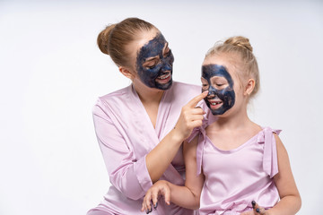 A young mother and a young daughter are sitting next to her in face masks. Facial skin care.
