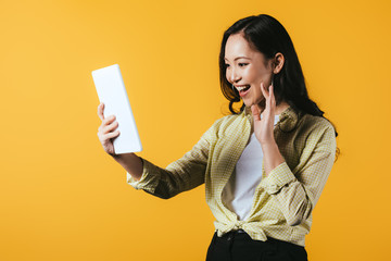 smiling asian girl waving and making video call on digital tablet, isolated on yellow