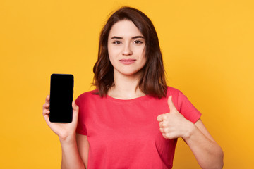 Close up portrait of beautiful woman holding phone with blank screen and showing ok sign with other hand, girl has dark hair, wearing red casual t shirt, posing isolated over yellow studio background.