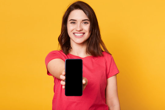 Cheerful energetic black haired female reaching out arm, holding smartphone in one hand, smiling sincerely, looking directly at camera, being in high spirits, wearing casual bright red t shirt.