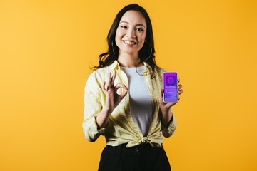 smiling asian girl showing ok sign and smartphone with shopping app, isolated on yellow