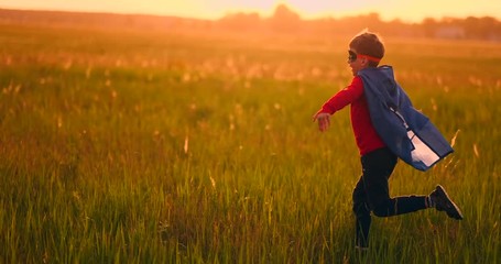A boy in a suit and a superhero mask running across the field at sunset on the grass