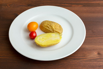 Slices of boiled potatoes and two tomatoes lie on a white round plate on a wooden brown table.