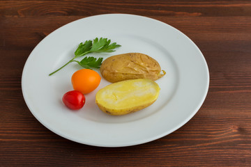 Slices of boiled potatoes, two tomatoes and a leaf of parsley, lie on a white round plate on a wooden brown table.
