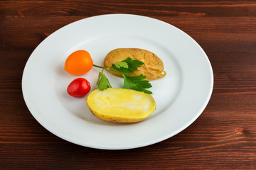 Slices of boiled potatoes, two tomatoes and a leaf of parsley, lie on a white round plate on a wooden brown table.
