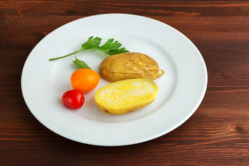 Slices of boiled potatoes, two tomatoes and a leaf of parsley, lie on a white round plate on a wooden brown table.