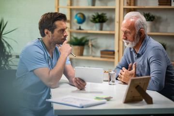 Serious doctor and senior man communicating while using touchpad during medical appointment.