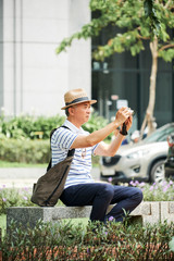 Asian tourist in hat and casual wear sitting on bench and taking photo on digital camera while sitting on bench in the city