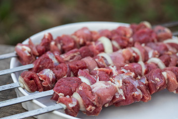 Five skewers with pieces of raw lamb with salt, black pepper and onion on a white round dish on an old wooden table, close up