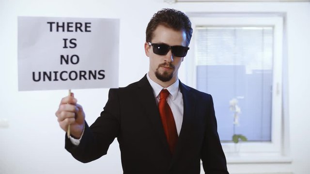Attractive man hold a sign There Is No Unicorns 4K. Medium shot of a male person in focus dressed up nicely with a red tie. Wall with clock and window in the background.