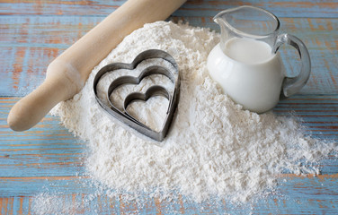 Flour, rolling pin and three baking hearts and a glass jug with milk among white wheat flour on blue wooden table boards