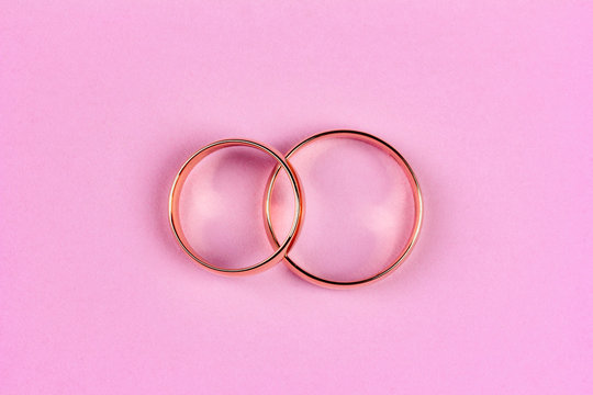 A Pair Of Gold Wedding Rings On A Pink Background, Top View Flat Lay