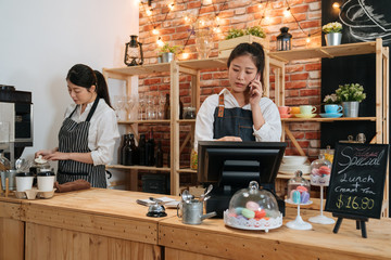 smiling waitress taking order on smartphone and using tablet in cafeteria. coffee shop staff talking online with customer while barista preparing beverage. two girl colleagues work together in cafe