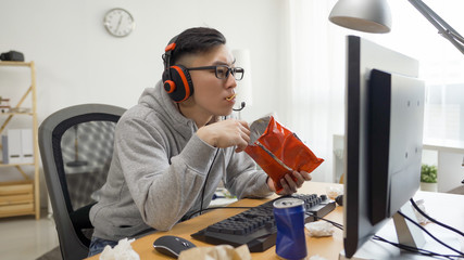funny asian man with headphones plays video games on computer and eats snack from pack. japanese otaku gamer in glasses enjoy chips while watching anime cartoon movie on pc screen on summer break. - obrazy, fototapety, plakaty