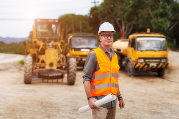 Handsome engineers wear protective helmets and are standing with road construction vehicles...
