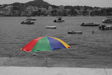 A black and white image of boats on the water with a multi-coloured sun umbrella in the foreground.