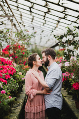 Young loving cheerful caucasian couple in glass greenhouse among colorful azalea flowers. Happy man and woman hugging and holding hands, spending time togehter.