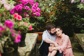 Fototapeta premium Young loving cheerful caucasian couple in glass greenhouse among colorful azalea flowers. Happy man and woman hugging and holding hands, spending time togehter.